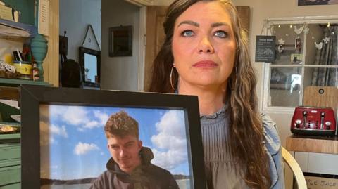 A woman with long dark hair, wearing a blue top and sat in her home, is holding up a picture of a young man with dark hair and a black hoody