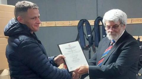 A man in a navy coat receiving a certificate from a man in a black suit with a red and blue tie.