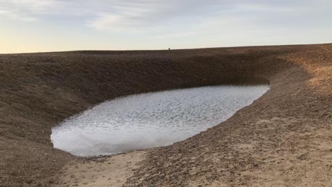 A picture of the hole on Felixstowe beach. A small body of water sits at the bottom of it and large shingle banks surround it.