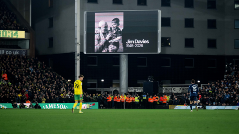 A black and white image of Jim Davies with a scarf round his neck holds a young boy in a football kit in his arms appears on a big screen above the pitch. To the left of the photo is a timer that says 80:34. Two players are on the pitch and match officials in orange and yellow high vis jackets stand underneath the screen and the stands are full.