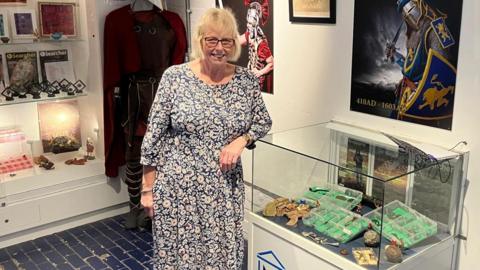 Lorraine is wearing a blue and white paisley dress and is leaning on a display cabinet that has some of her finds inside. She is wearing glasses and is smiling at the camera.