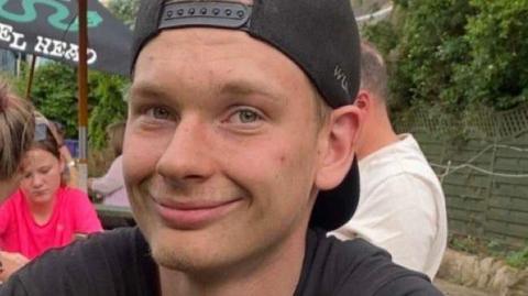 Logan Parling is smiling at the camera. He is wearing a backwards black baseball cap and has a black T-shirt on. It looks like he is sitting in a pub garden and there are other people in the background and a parasol on an outside table