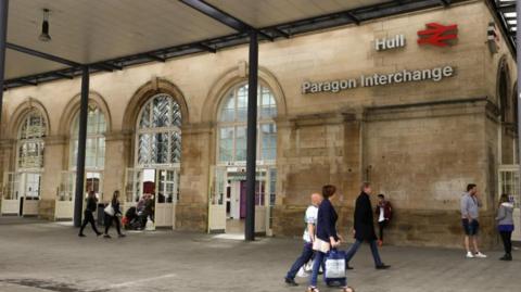 About 10 people walk across a wide paved area underneath a wooden roof supported by two metal pillars. They are approaching the entrance of a railway station which is built from stone and has four archways containing double doors with arched windows above. The words Hull Paragon Interchange in white lettering and a red British Rail double arrow symbol are affixed high on the stone wall.