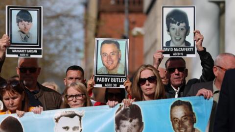 A shot of a number of family members of those killed, they are standing behind a banner which has a blue background and the faces of some of their loved ones. Three placards are being held up - of Margaret Gargan, Paddy Butler and David McCafferty - old photos with the ages they were when they died.