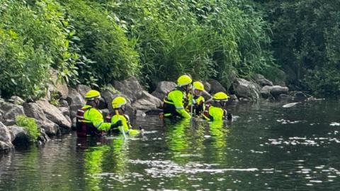 Five people in high-vis outfits and helmets waist deep in a river. Behind them to the left is the river bank, which is covered in green foliage.