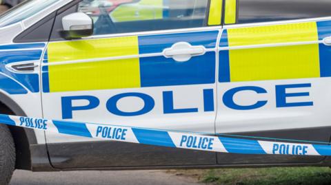 A stock image showing a police car with blue and yellow hi-vis decals, parked behind blue and white police cordon tape at the scene of a collision.
