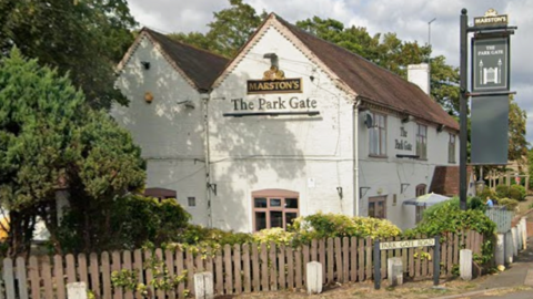 A white-walled building stands behind a wooden fence with the words "The Park Gate" on its side while a pub sign hangs in front of it.