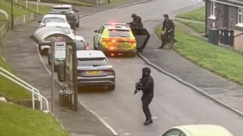 Armed police officers dressed in all black on a residential street. A police car with red lights flashing can also be seen.