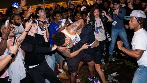 A crowd of people gather are two men fighting in the street 