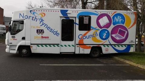 A mobile library, with a central door. It is white, with coloured logos reading: "North Tyneside Council", "I'm the plus bus" and images of an open book and a lap top in purple circles.