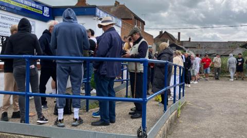 A long line of between 30 and 40 people queuing at the entrance to Southend United's ticket office, which is a small blue and white building. Outside it is a gravel car park with the queue winding through it.