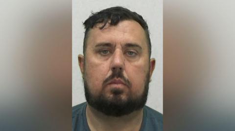 Mugshot of Mark Twizell. He has a short black hair and beard and a long round face.