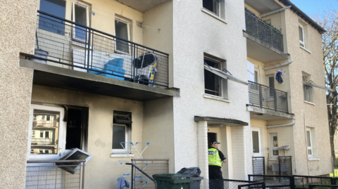 A view of the Niddrie Mill flat with black fire damaged windows. There are blocks of flats with white and light stone fronts. Each has a balcony in between the blocks. 