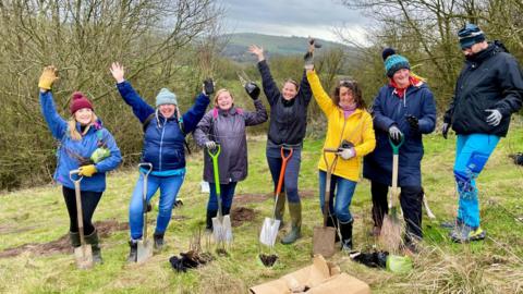 A group of young adults, mostly women, with shovels dressed in winter clothing planting trees in open land surrounded by forest. 