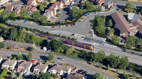 A CGI image from above of a planned railway station in a residential area. The train line runs between the houses and a purple and orange train is on the track. There is also a car park behind the railway line.