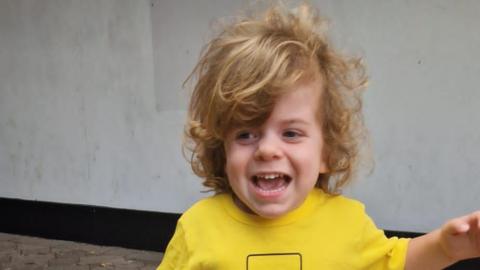 A photo of Wilfred. He is smiling and looking away from the camera. He has long, curly blonde hair and is wearing a yellow T-shirt. He is holding one arm away from his body.