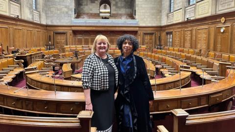 Two women stand in a council chambers. One woman is white and blonde and wearing a black and white dress suit. The other woman in black with curly dark hair. She is wearing a blue and black dress.