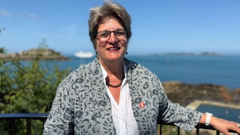 Deputy Jayne Ozanne is wearing a jacket with small navy flowers against a paler blue background, and a white shirt. She is standing against a blue rail with a bright blue sea behind her. She is wearing a black beaded necklace and large black framed glasses.