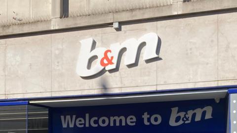 A white and orange B&M sign with signage below. On a stone wall. 