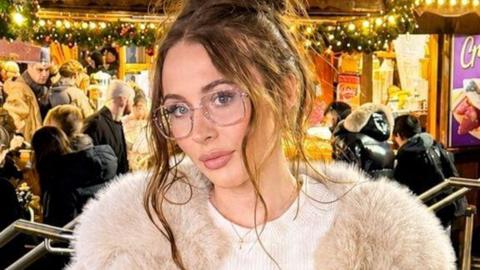 A woman with wavy brown hair which is tied up. She is wearing glasses and a fluffy beige jacket. She appears to be standing in a pub