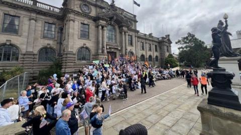 A crowd of people outside South Shields Town Hall, a large stone building in the Edwardian Baroque style. There are columns on either side of the doorway, which is topped by a large pediment and a flag pole. The ground floor features a row of arched windows, and the upper floor square ones. The crowd, standing on the steps leading towards the entrance, and round to the side, are holding flags and banners.