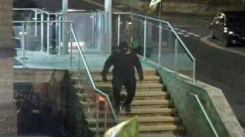 A man in all black clothing walking down some stone steps with glass barriers either side.