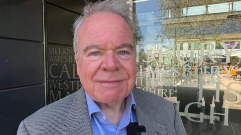 Man in a blue shirt and a grey suit jacket smiling at the camera. He is standing in front of an office building. Places in Wiltshire are written on the glass doors.