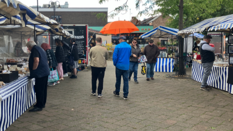 Blue and white-striped market stalls either side of a paved area with people walking towards and away 