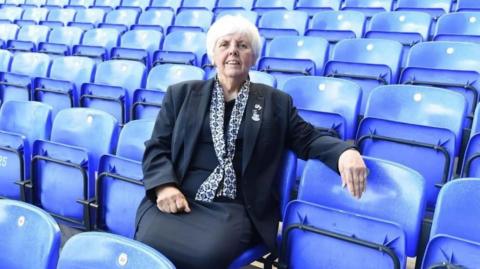 Lily Barnes is photographed sitting in one of Goodison Park's stands and smiling at the camera. There are rows of blue seats  all around her.