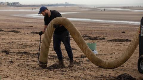 A man is on a beach, holding a large plastic hose. He's dressed in black, with a navy blue cap
