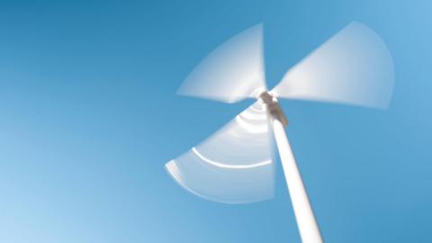 A view from the ground of a white wind turbine, its three blades blurred as they spin against a clear blue sky.