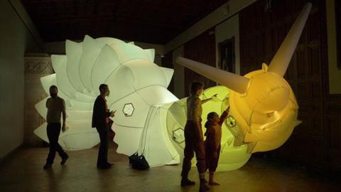 Huge illuminated inflatable snail looms above four visitors in a darkened museum hall as a child reaches out to touch the yellow head. 