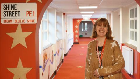 Becky Waters is wearing a brown  cardigan with stars on it, she is standing in the school corridor underneath an archway, that is painted red with gold stars. On the archway is a sign saying "Shine Street PE1".