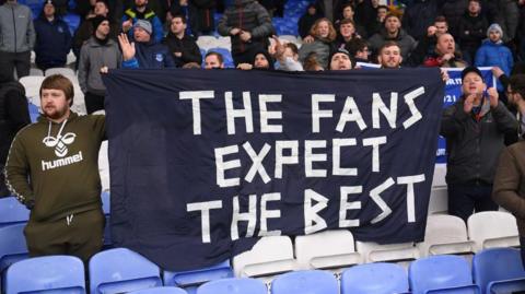 Fans hold a bedsheet banner saying 'The Fans Expect The Best' in a football stadium.