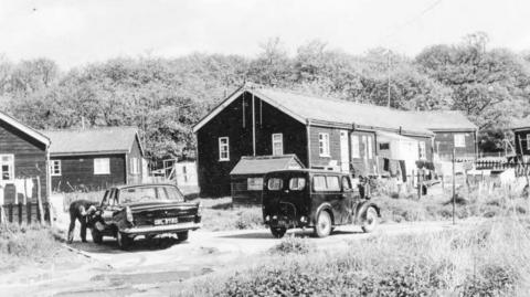 An archive black and white image from Slade Camp when residents still lived there. There are houses and cars parked by them. A man can be seen working on a car.