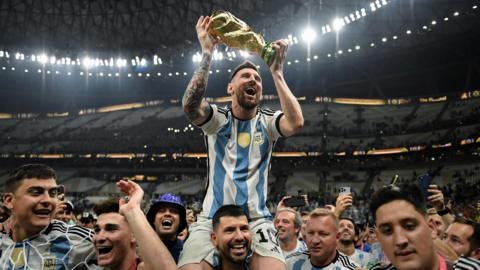 Lionel Messi lifts the World Cup aloft whilst being carried on the shoulder of countryman Sergio Aguero inside a football stadium. They are surrounded by others celebrating Argentina's win and filming on their mobile phones.