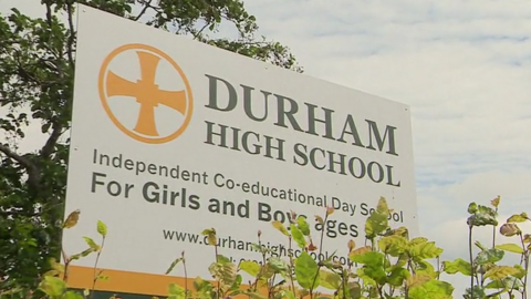White sign with a yellow circular emblem. Black text which says Durham High School. Green trees are in the back left and front right, slightly covering the sign. 