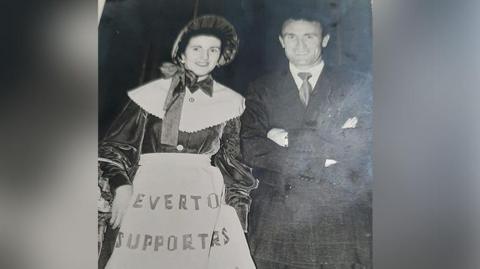Everton FC toffee lady Mary Morgan (nee Gorry) wearing Victorian style confectionary attire with white pinny and bonnet with Tommy Eglington who played for Everton wearing a suit taken between 1953 and 1956.  