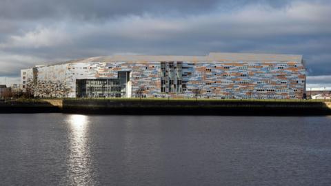 A general view of Middlesbrough College taken across the dock, with the water in the foreground. The large building is clad with grey, blue and orange material creating an irregular pattern.