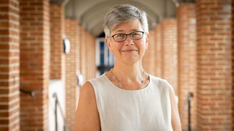 A woman with short grey hair and glasses. In the background is a brick walkway. 