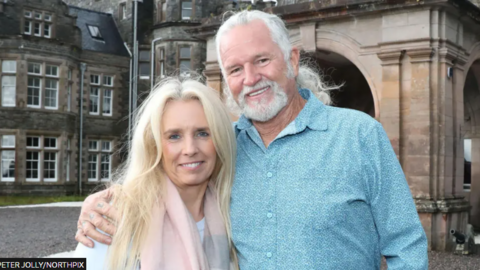 Suzanne agus Lance Holmquist. Boireannach le falt fada bàn. Tha stoc pinc mu a h-amhaich. Fireannach ri a taobh le falt fada liath ceangailte agus feusag. Tha lèine ghorm. Tha iad nan seasamh air beulaibh caisteil.