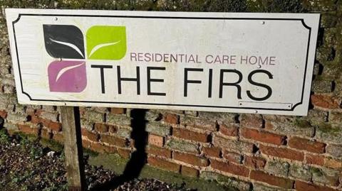 A white rectangular sign, with a black, green and purple leaf motif, saying "Residential Care Home The Firs". It is next to a brick wall.