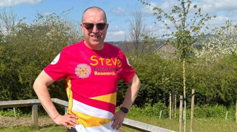 Stephen Rhodes is standing in his garden with young trees behind him. He is a 55 year old man wearing sunglasses and a red, yellow and white top with the word Steve and Brain Tumour Research printed on it