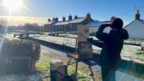 A man in a black wooly hat, gloves, coat and trousers is painting on an easel by a snowy canal loch. The sun is beaming in the top left of the image. The man is sheilding his eyes with his left hand and painting with the other. He has a pallette of coloured paint and bags beside his easel. Terraced houses run alongside the canal.