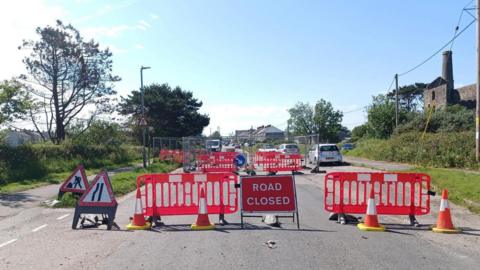 Red barriers, a Road Closed sign and traffic cones are in the foreground with metal fencing set up as a barrier around holes further down the road where there are more barriers. There are mine buildings with a tower on the right, close to the road.