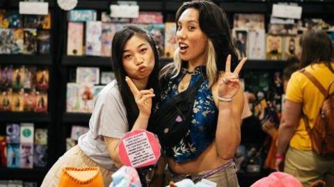 Two young Japanese women - one in a grey t-shirt and yellow dress, the other in a traditional Japanese blue dress with patterned flowers. They are posing for the camera.