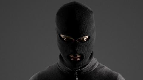 A person stares at the camera while wearing a full-face balaclava and a zipped up, grey jumper. They are standing in front of a dark grey wall.