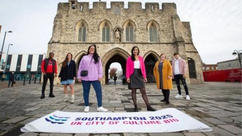 A group of six people standing in front of Southampton's Bargate, with a banner on the ground saying 'Southampton 2025 UK City of Culture bid'