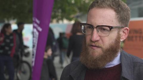Daniel Randall in a red jumper and grey jacket. He has glasses, a beard and one earring showing. 