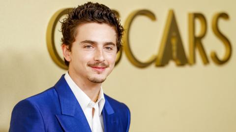 Timothee Chalamet in a bright blue suit and white shirt, posing in front of the word Oscars on a wall in large gold letters, at the 98th Oscars Nominees Luncheon at the Beverly Hilton Hotel on 10 February 2026